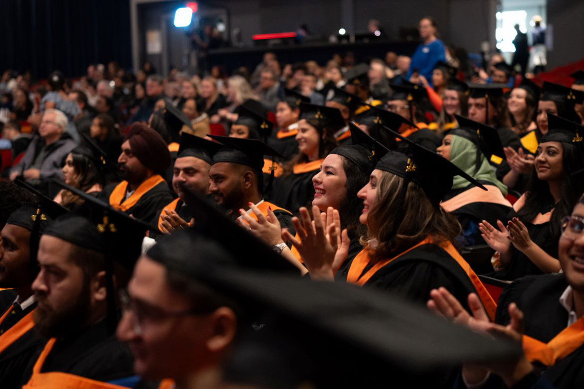 Graduates sitting during convocation ceremony, in academic regalia, with ambassador in the background in a blue gown