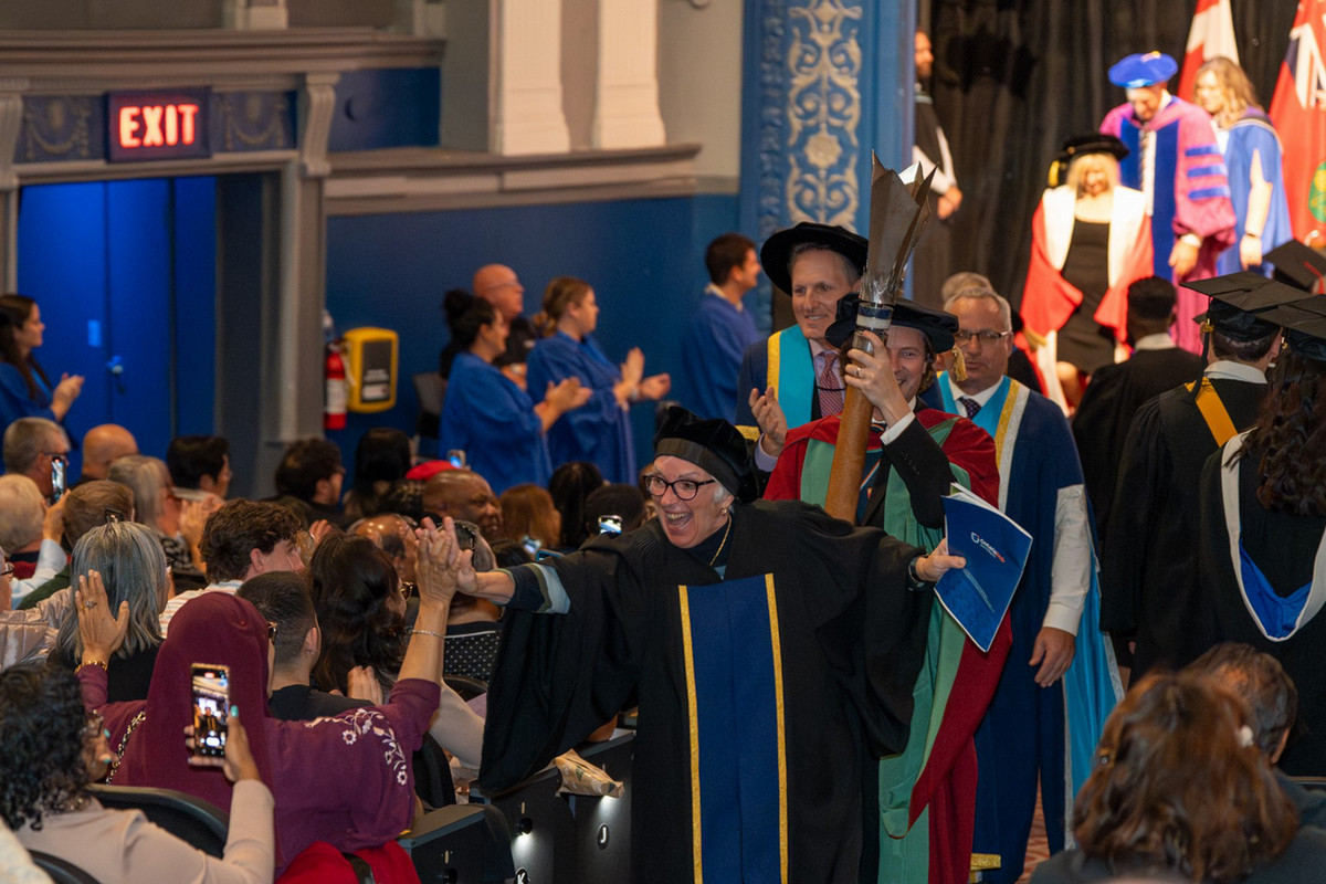 Convocation celebration photo while one faculty member high-fives attendees, smiling broadly. The background shows additional faculty and ambassadors in academic robes.