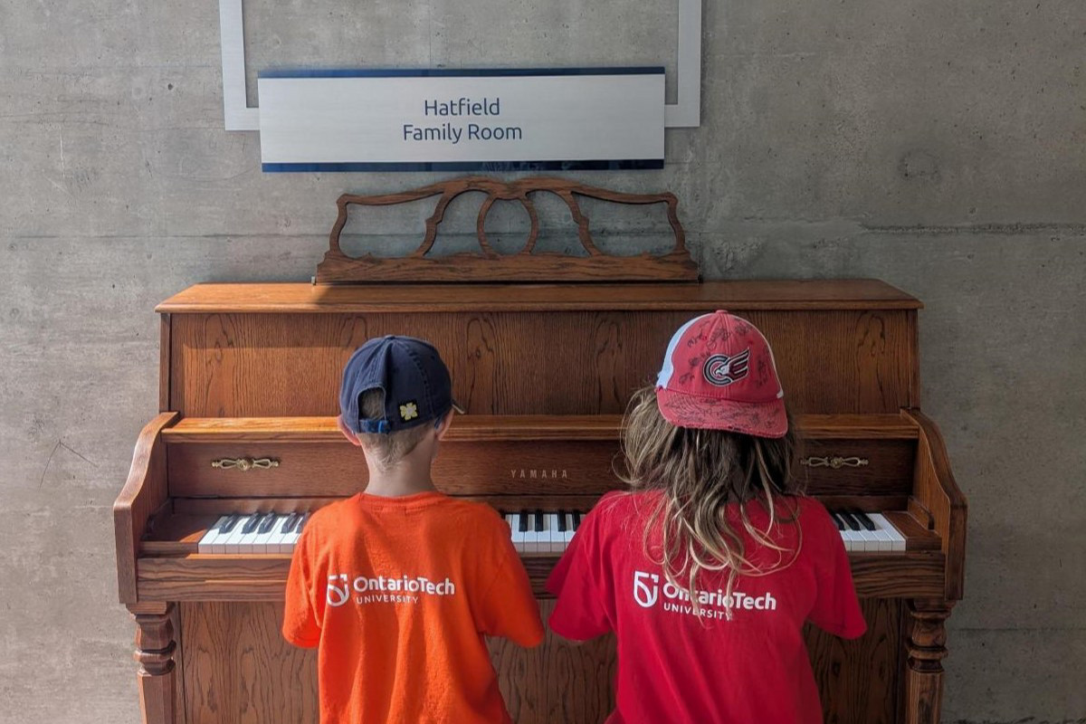 Two children playing a piano and wearing t-shirts that have Ontario Tech University Logo.