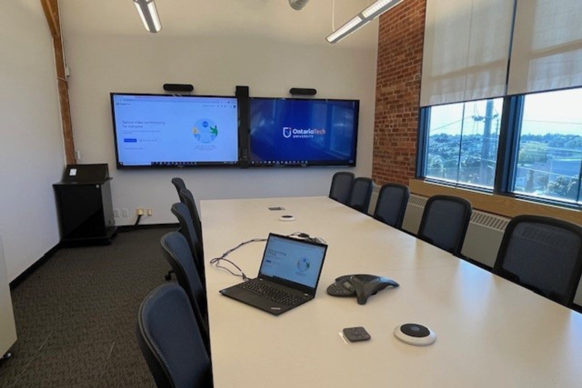 A modern conference room with a long white table, chairs, and a laptop connected to two wall screens showing a Google Meet and Ontario Tech logo. Large windows show a view of downtown Oshawa.