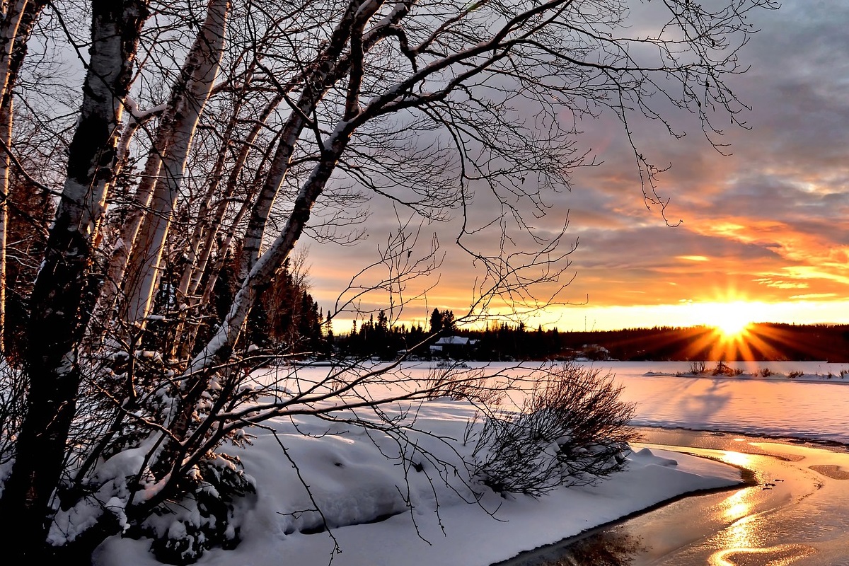 Sun setting over winter landscape including leafless trees, snow on the ground and a small stream.